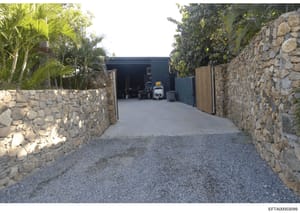 Photograph of a residential property's exterior showing a gated driveway entrance flanked by stone walls, leading to a dark-colored metal outbuilding or garage structure. The image captures the property's access point and security features, with trop