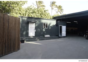 This is an evidence photograph showing the exterior of a modern, dark-colored modular or prefabricated building structure. The building features emergency exit signage visible on a white door, metal framework, and is situated in a tropical setting wi