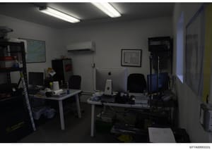 This is an evidence photograph showing an interior office or workspace with multiple computer workstations, including Mac computers, desks, chairs, shelving, and various office equipment. The image appears to be from an investigative search or proper