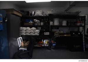 This photograph shows an interior storage or utility space containing industrial shelving units stocked with various household and maintenance supplies, tools, and materials. The image appears to be evidence documentation from a property search or in