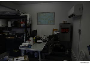 This photograph shows the interior of an office or administrative workspace, featuring a desk workstation with computer equipment, storage shelving, and various office supplies. The room contains a wall-mounted map, air conditioning unit, and a 'KEEP