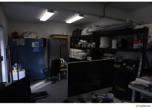 This is a photograph of what appears to be a maintenance, storage, or utility room. The space contains various equipment including refrigeration units, shelving with supplies, cleaning materials, and office furniture. The room is well-lit with fluore