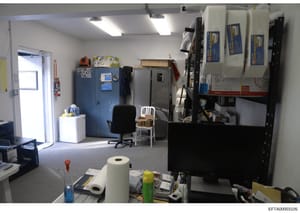 This is a photograph of an interior workspace showing office furniture, storage cabinets, equipment, and supplies. The room appears to be a maintenance, storage, or administrative area with fluorescent lighting, blue storage units, shelving with vari