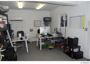 This is a photograph of an office or workspace interior, showing computer stations, storage equipment, and various office supplies. The room appears to be used for administrative or technical work, with multiple desks, an iMac computer, printers, and