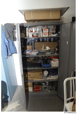 This photograph documents the contents of a large storage cabinet or closet, showing various electronic equipment, boxes of supplies, tools, and personal items. The image appears to be evidence documentation from a property search or inventory, showi