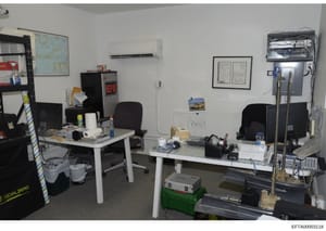 This is a photograph of an office or workspace interior containing two white desks with black office chairs, computer equipment, filing systems, and various office supplies. The space appears to be used for administrative or operational purposes and 
