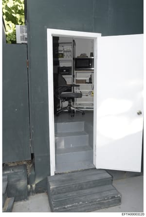 This photograph shows an interior equipment room or closet containing electrical infrastructure and computer equipment. The image captures a white-framed doorway opening into a narrow space with an electrical panel mounted on the upper wall, a black 