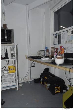 This is an evidence photograph showing an interior office or workspace, likely from one of the properties involved in the Epstein investigation. The image depicts a functional work area with communication devices, computer equipment, shelving, and to
