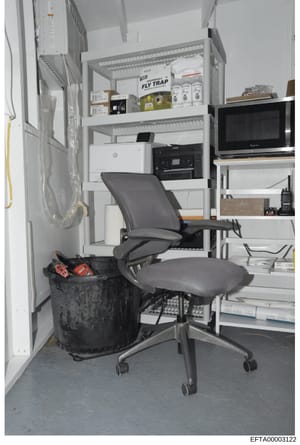 This is a photograph of a small office or work room containing shelving units with various items including office equipment (HP printer, multifunction devices), pest control products (Fly Trap boxes), a microwave, electronics, and miscellaneous suppl