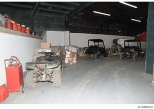 This photograph shows the interior of a large industrial storage facility or warehouse containing multiple all-terrain vehicles (ATVs), utility carts, and various stored equipment. The space includes red fuel containers, pallets of supplies, and main