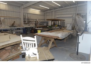 This photograph shows a large metal-frame workshop space containing woodworking equipment, materials, and furniture in various stages of completion. The space includes wooden workbenches, a white chair, lumber, power tools, a large industrial fan, an
