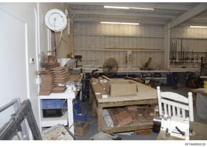This photograph depicts an industrial workshop or storage space containing various woodworking materials, tools, and equipment. The image shows shelving units, stacked wooden pieces, workbenches, hand tools, and mechanical equipment arranged througho