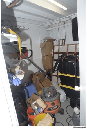 This photograph shows the interior of a storage area or utility room containing various tools, equipment, and household items. The space includes a RIDGID wet/dry vacuum, power tools, extension cords, cardboard boxes, a bed frame, and other miscellan