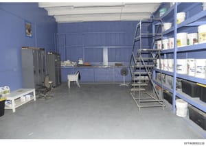 This photograph depicts an industrial storage or warehouse room with bright blue walls and concrete flooring. The space contains metal shelving units stocked with supplies (white buckets and containers), HVAC equipment, office furniture including cha