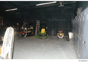 This photograph shows the interior of a storage garage or equipment shed containing various maintenance and landscaping equipment. The space includes a green John Deere zero-turn radius lawn mower, a smaller yellow and red mower, ladders, tools, and 