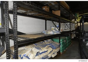 This photograph documents the interior of a warehouse or storage facility containing multiple shelves with packaged materials in blue and tan/beige colored bags. The image shows industrial metal shelving units with various containers and supplies sto