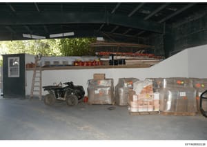 This photograph shows the interior of a warehouse or storage facility containing multiple palletized items wrapped in plastic with inventory tags, a black ATV/four-wheeler vehicle, construction materials, and storage shelving. The image appears to be