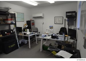 This photograph documents an office interior containing two workstations with desks, office chairs, storage shelving, and various equipment. The space appears to be used for administrative or operational purposes, with a wall-mounted map, calendar, f