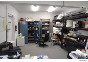 This is a photograph of a storage and maintenance room within an Epstein property, showing organized tool storage, cleaning supplies, and household maintenance equipment. The room contains blue metal tool cabinets, shelving units with supplies, a des