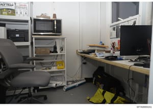 This is a photograph of a small office or workspace containing computer equipment, shelving, a desk, and various storage items. The room appears to be a work area with a microwave, multiple monitors, filing materials, and office supplies. The photo w