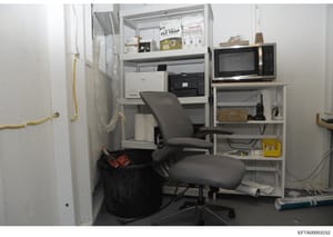 This photograph shows a small, cramped office or utility room with minimal furnishings and equipment. The space contains a desk setup with computer equipment, shelving units, and basic office supplies. The room appears to be part of an Epstein proper