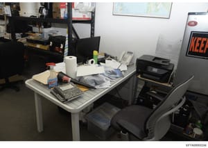 This is an evidence photograph of an office workspace showing a desk with various items including computer equipment, papers, office supplies, and personal effects. The cluttered desk and surrounding area suggest this may be documentation of a locati