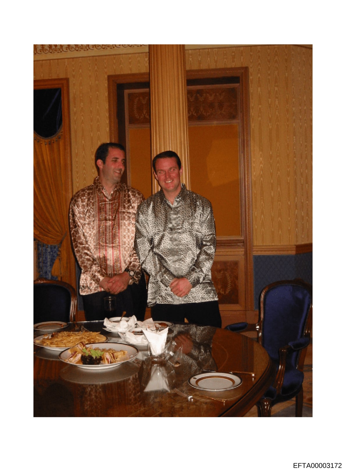 This photograph shows two men in patterned jackets standing in an elegantly decorated formal room with damask wallpaper, wood paneling, and classical architectural details. A dining table with place settings and food is visible in the foreground. The