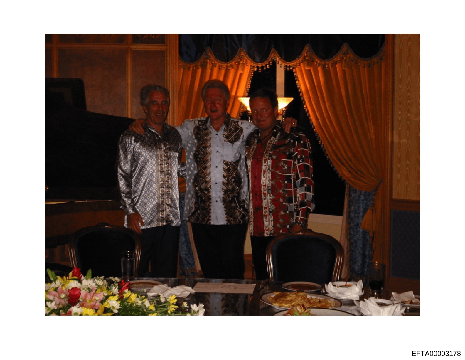 A color photograph showing three adult men wearing distinctive patterned and ornate jackets at what appears to be a formal indoor event or dinner. The setting features elegant furnishings including orange velvet curtains with decorative trim, wooden