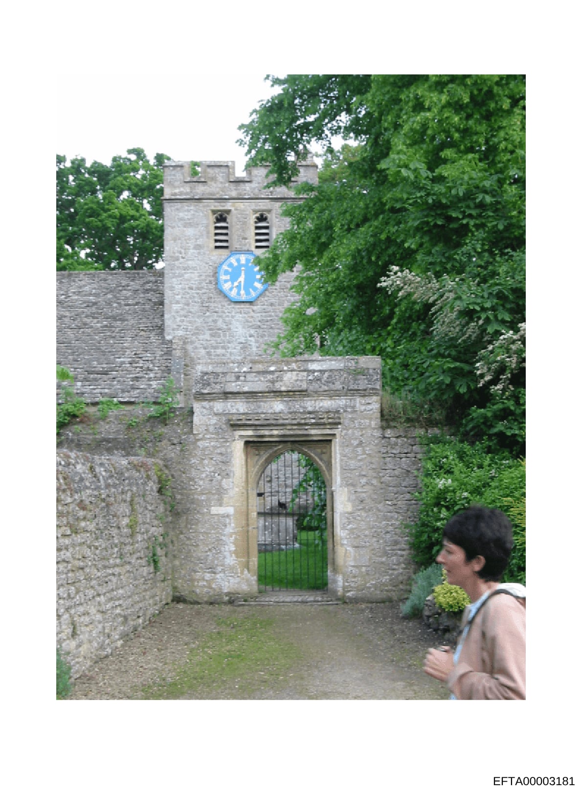 This is a photograph of a historic stone church or fortified religious structure featuring a crenellated tower with a blue clock face. The image shows the building's gated entrance with a Gothic-style arch and heavy wooden door with metal bars. The s