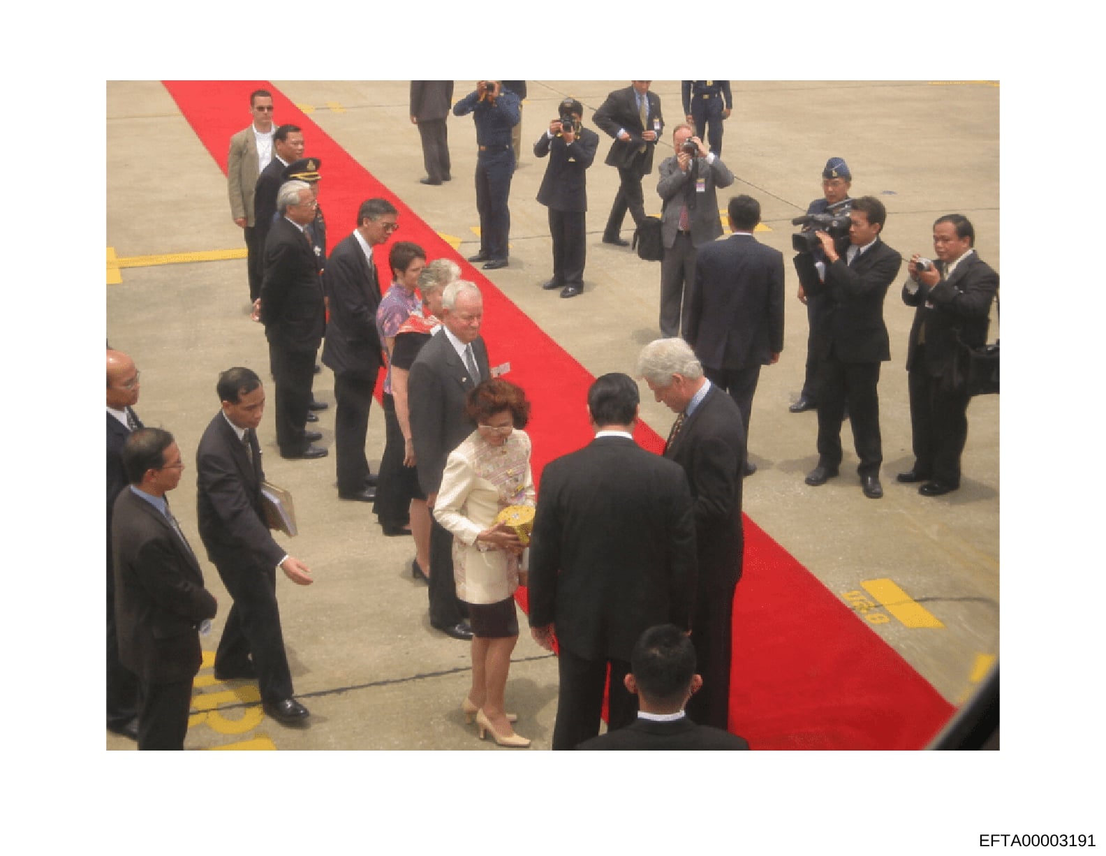 This photograph shows a formal diplomatic or state reception on an airport tarmac, featuring numerous officials in business attire gathered around a red carpet. The scene includes security personnel, media with cameras, and what appears to be a digni