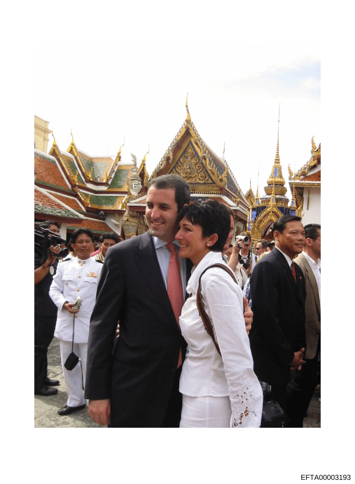 This photograph documents an official visit to a Thai Buddhist temple, showing a man in a dark suit with red tie and a woman in white clothing walking together in the temple courtyard. The image appears to be from a formal diplomatic or official stat