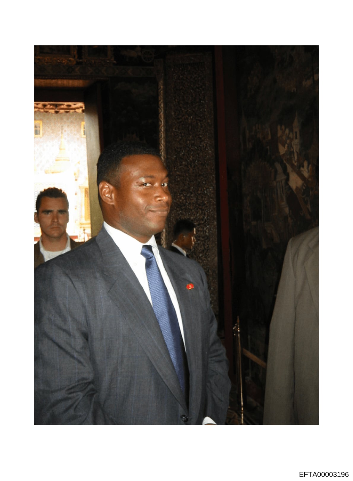 This is a photograph of a man wearing a dark gray business suit with a blue tie and small red pin, photographed indoors at what appears to be a formal event or gathering. The image shows the subject in profile/three-quarter view against a dark textur