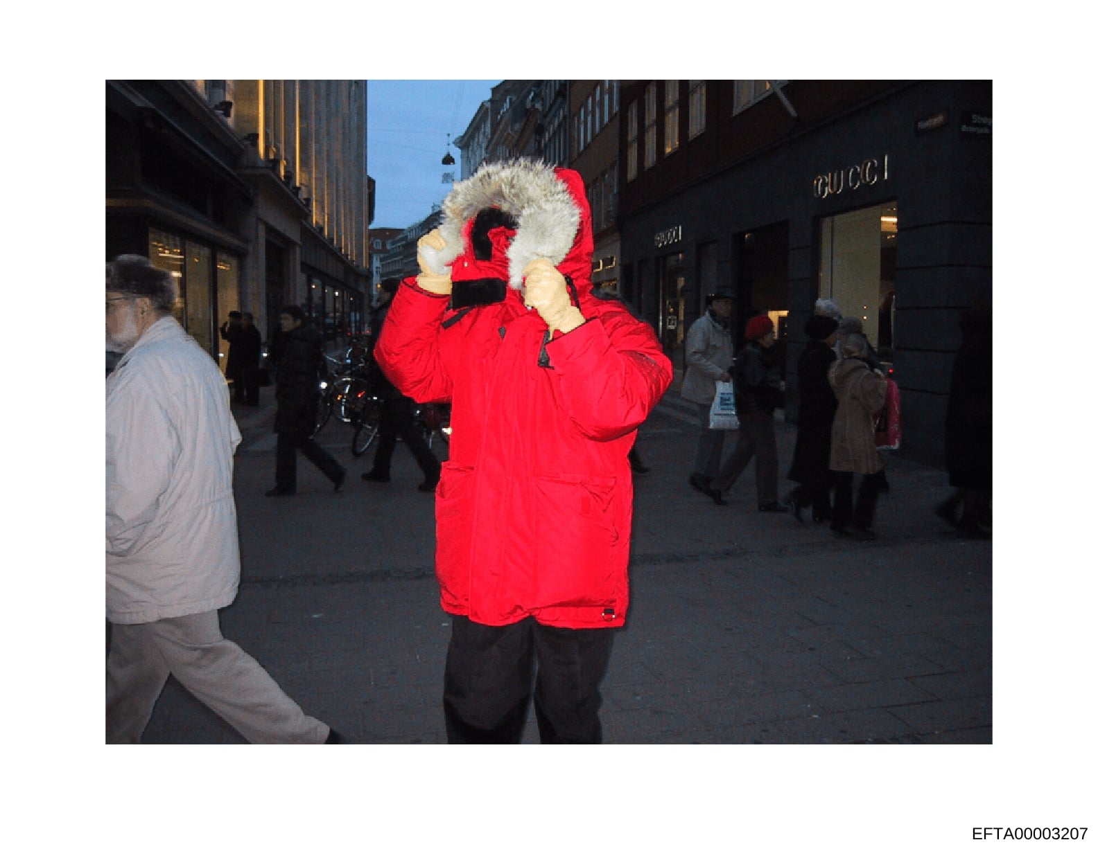 This is a photograph showing a person wearing a bright red winter parka with a large fur-lined hood on what appears to be a busy urban shopping street. The image captures a public street scene with Gucci storefront visible and various pedestrians. Th