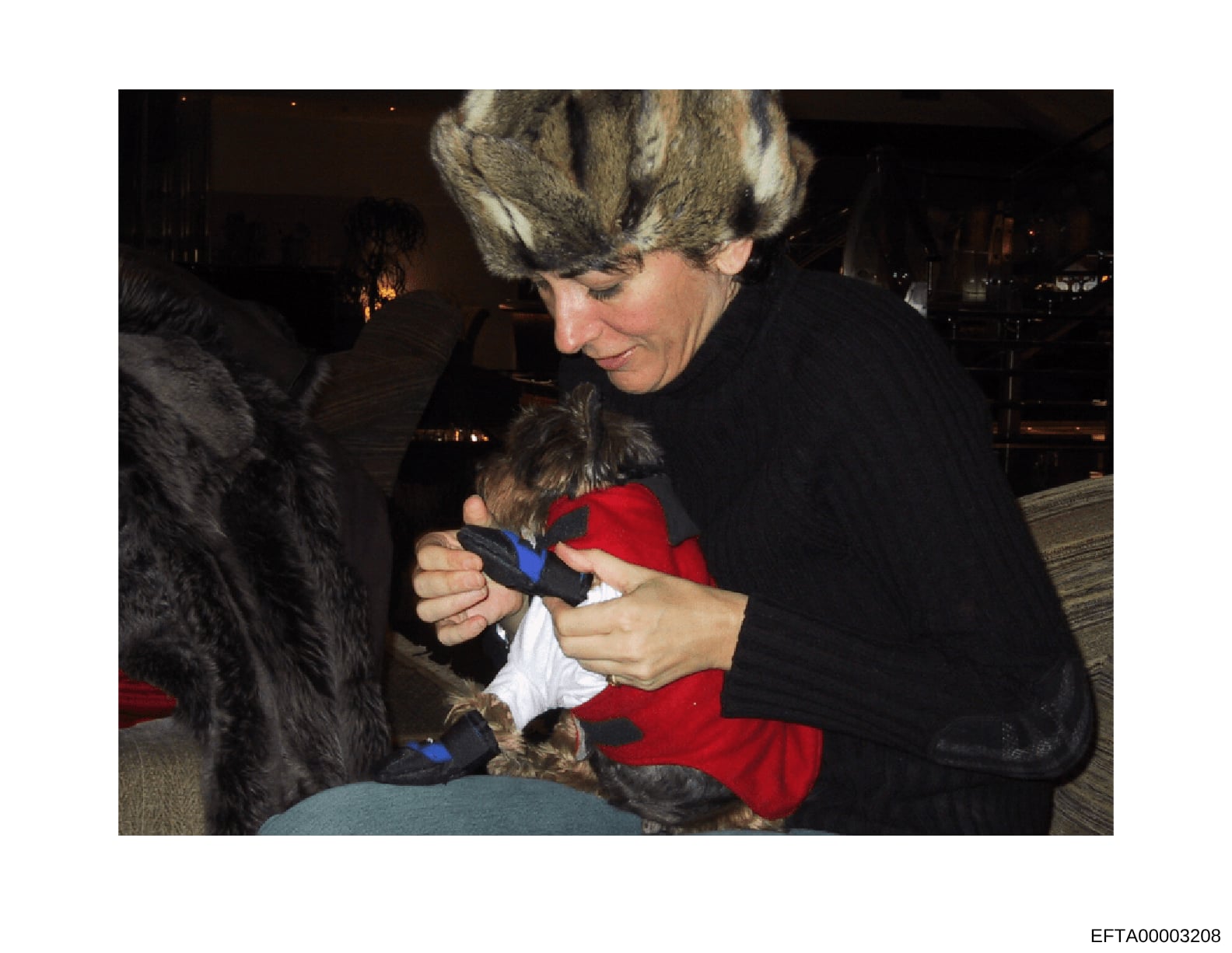 This is a photograph showing a woman wearing a fur hat and black coat holding a small dog dressed in red clothing at what appears to be an indoor social gathering. The image is included in the Epstein investigation files but lacks contextual informat