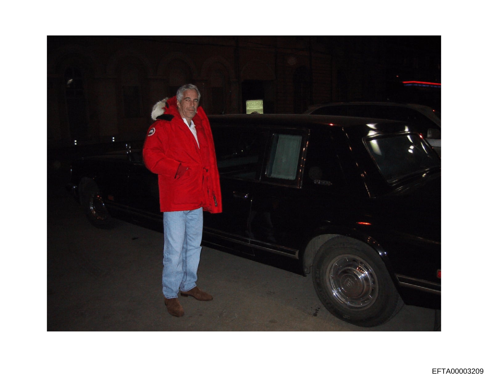This is an evidence photograph from the Epstein investigation files showing a man wearing a distinctive red jacket with fur collar and light blue jeans standing next to a black vehicle at night. The photo appears to be taken in a parking garage or si