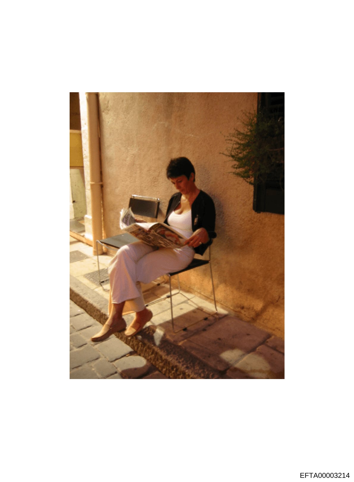 This is a color photograph showing a woman seated in a courtyard or alleyway area reading a magazine. She is dressed casually in a white top and pink/lavender pants with a black cardigan. The setting appears to be a Mediterranean or European-style re