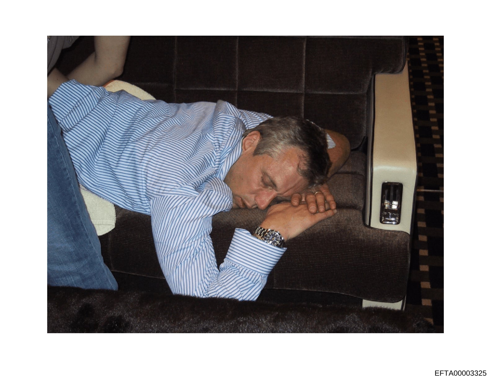 This is an evidence photograph showing a man wearing a blue and white striped shirt reclining or lying down inside what appears to be a vehicle with dark upholstered seating. The image appears to be from investigative documentation, with a visible Ba