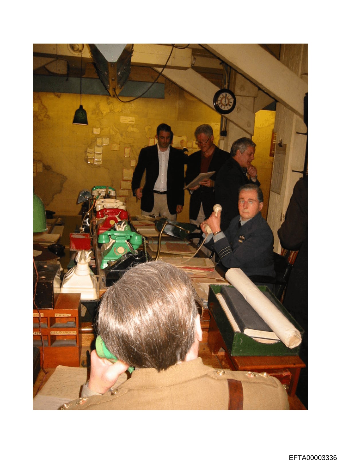 This photograph shows approximately five men in business attire gathered inside a basement or industrial space with yellow walls, examining what appears to be printing or manufacturing equipment. The image appears to be from an official tour, inspect