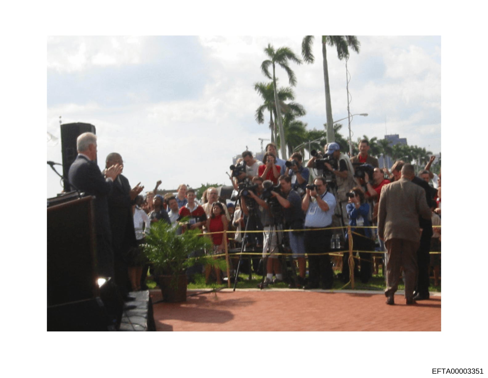 This photograph depicts a formal public event or ceremony taking place outdoors in a tropical setting, with numerous attendees gathered behind barriers and officials or speakers positioned at a podium. The image shows what appears to be an organized