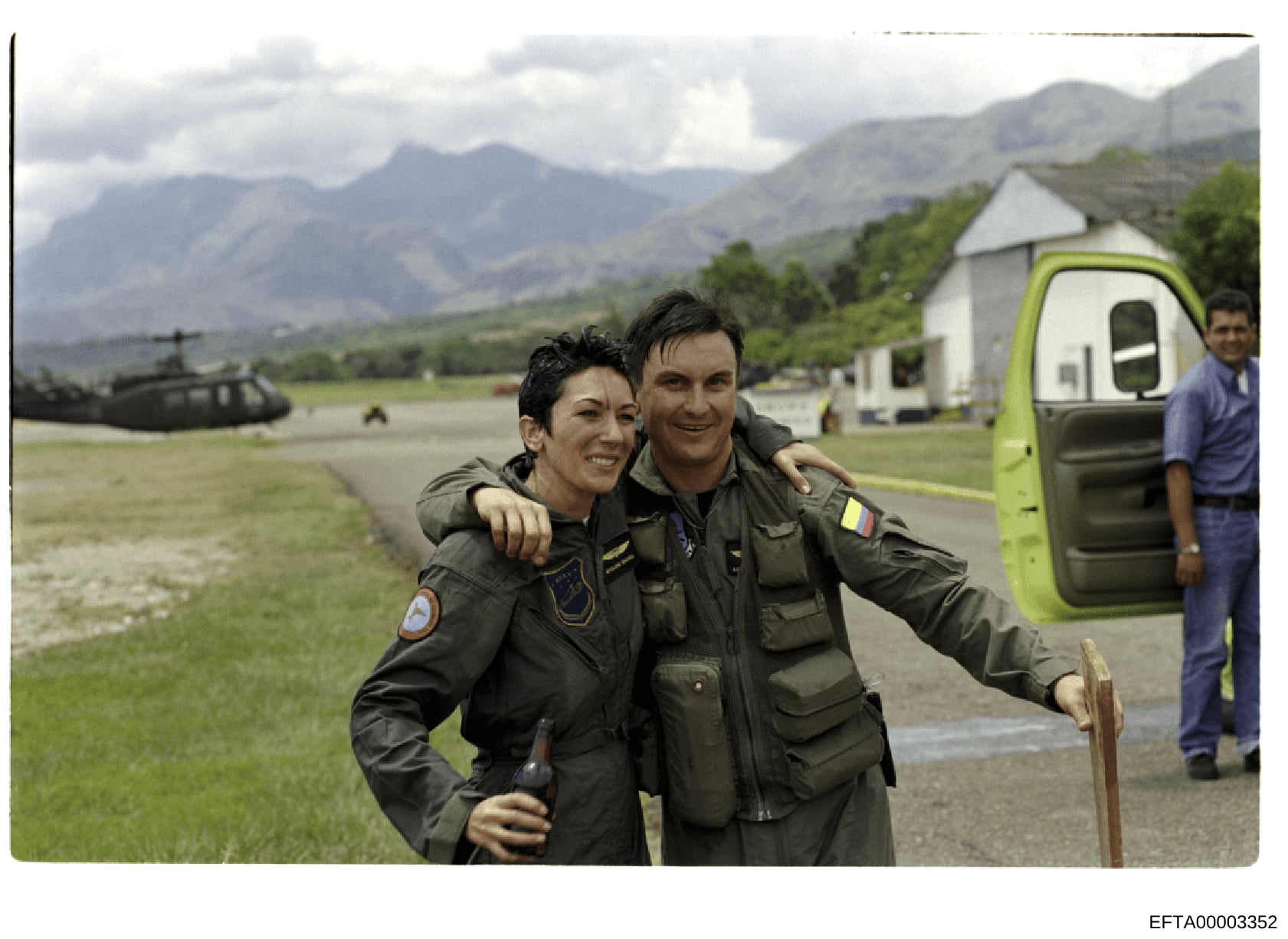 This photograph shows two military personnel in flight suits posing together at what appears to be a remote mountain airfield or landing zone. A military helicopter is visible in the background on the left, along with support buildings and a lime-gre