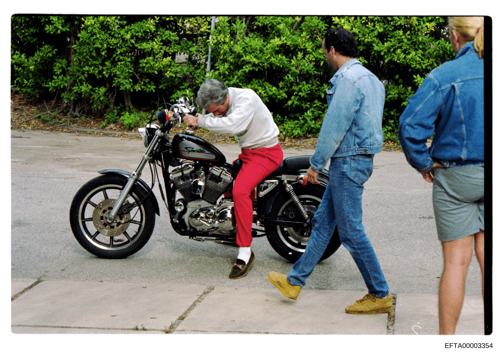 This is a color photograph showing a man with gray hair sitting on a motorcycle while three other people stand nearby observing. The image appears to be taken outdoors on a paved surface with green hedges in the background. The context and relevance 