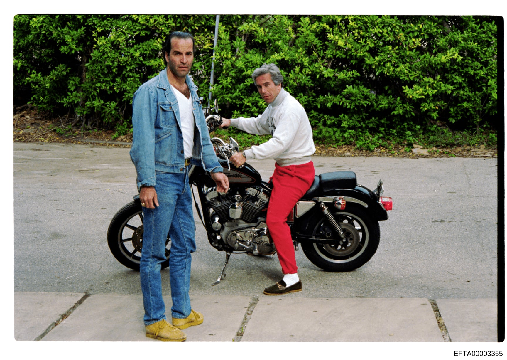 This is a photograph showing two adult men posed with a dark motorcycle in what appears to be a parking lot or paved outdoor area. The image appears to be a casual posed photo, likely from the 1970s or 1980s based on clothing styles and photograph qu