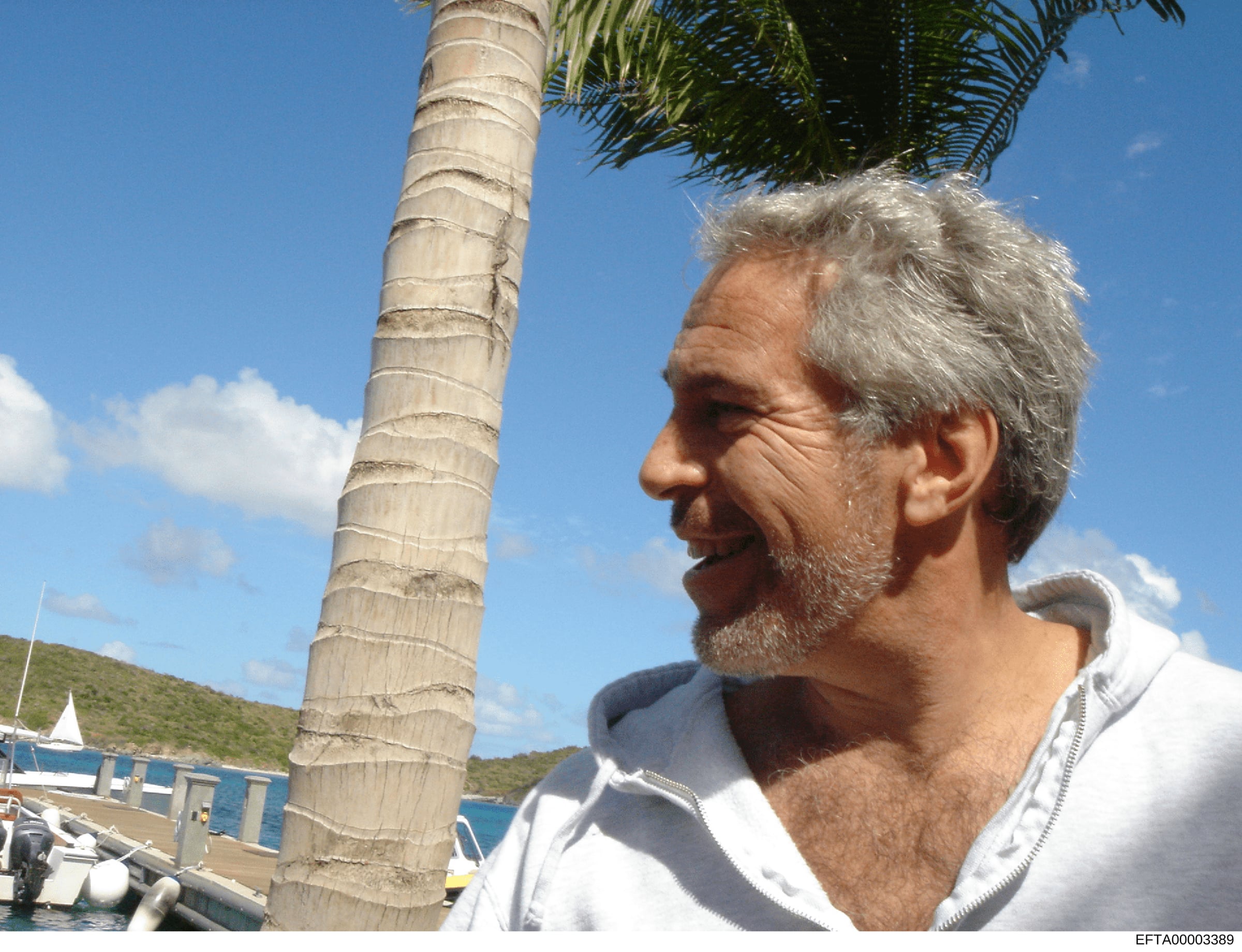 This is a candid photograph of a man with gray hair and facial hair at what appears to be a tropical marina or waterfront location. The image shows a person in profile wearing a white/light-colored hooded jacket, with palm trees, clear blue sky, boat
