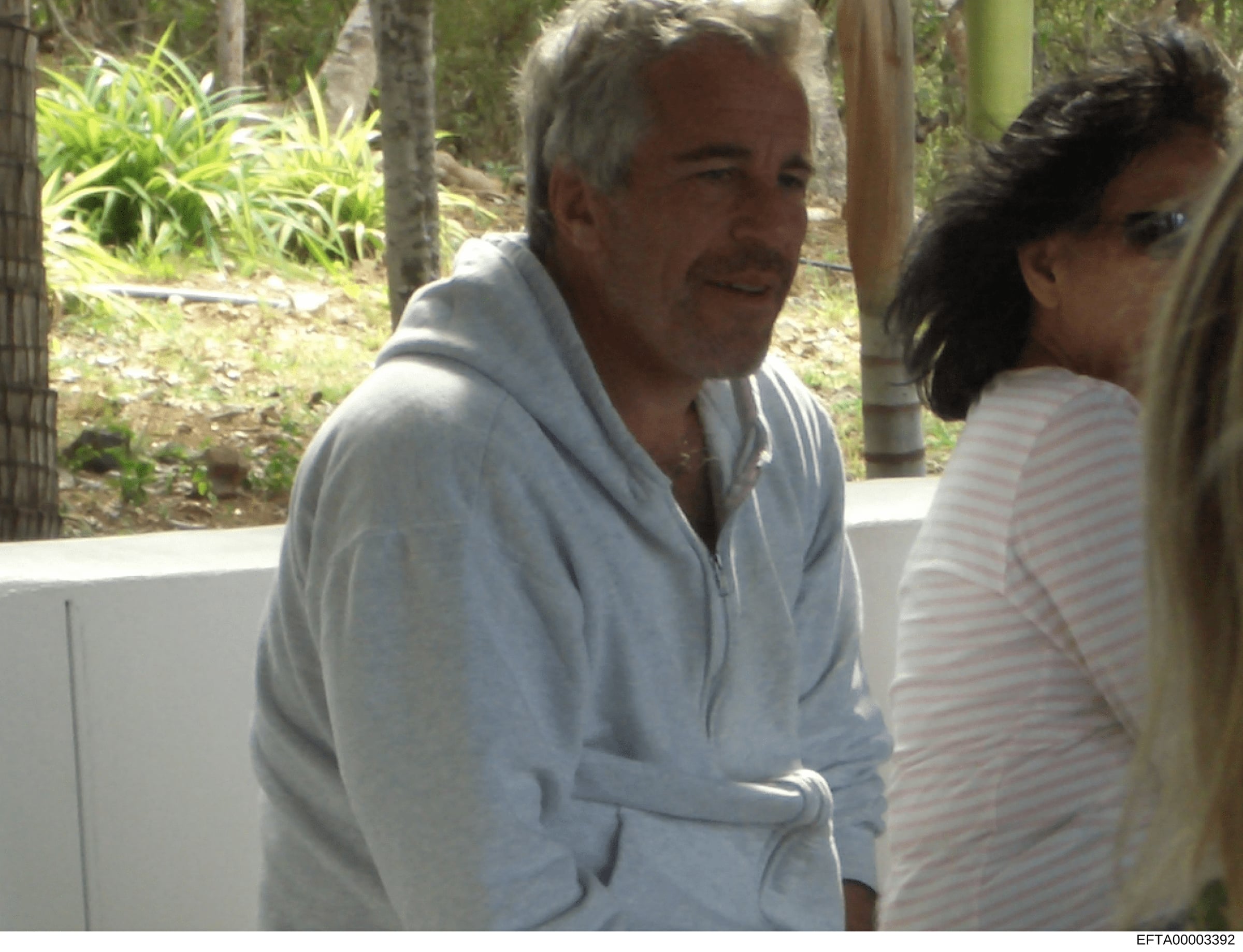 This is a photograph showing two people in an outdoor setting with tropical vegetation visible in the background. The image appears to be from investigative files and captures an informal moment at what looks like a residential property or estate. Th