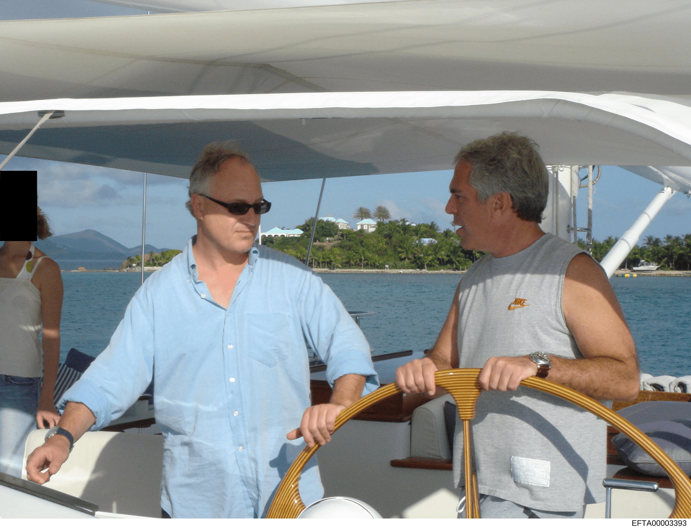 This photograph shows two men on the deck of a yacht in clear tropical waters, with coastal properties and mountains visible in the background. The image appears to be documentation of a social or leisure activity, potentially relevant to understandi