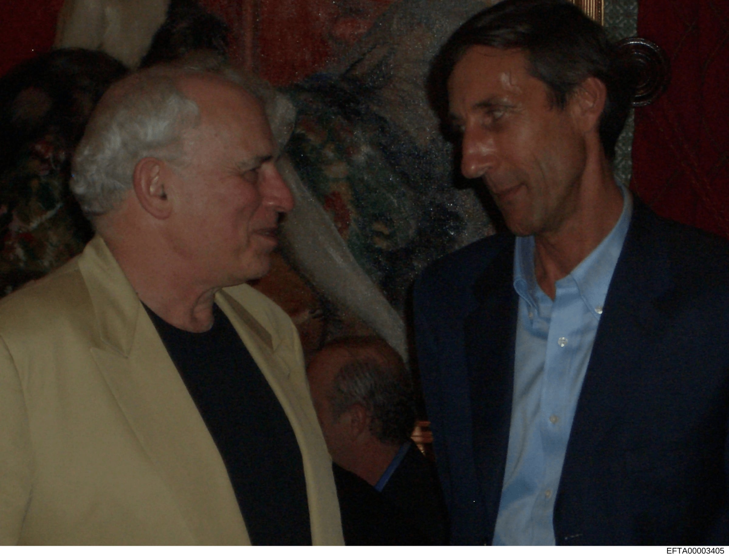 This photograph shows two men engaged in conversation at what appears to be an indoor social or professional event. The image quality and color palette suggest it was taken in the 1990s or early 2000s. One man wears a yellow blazer over dark clothing