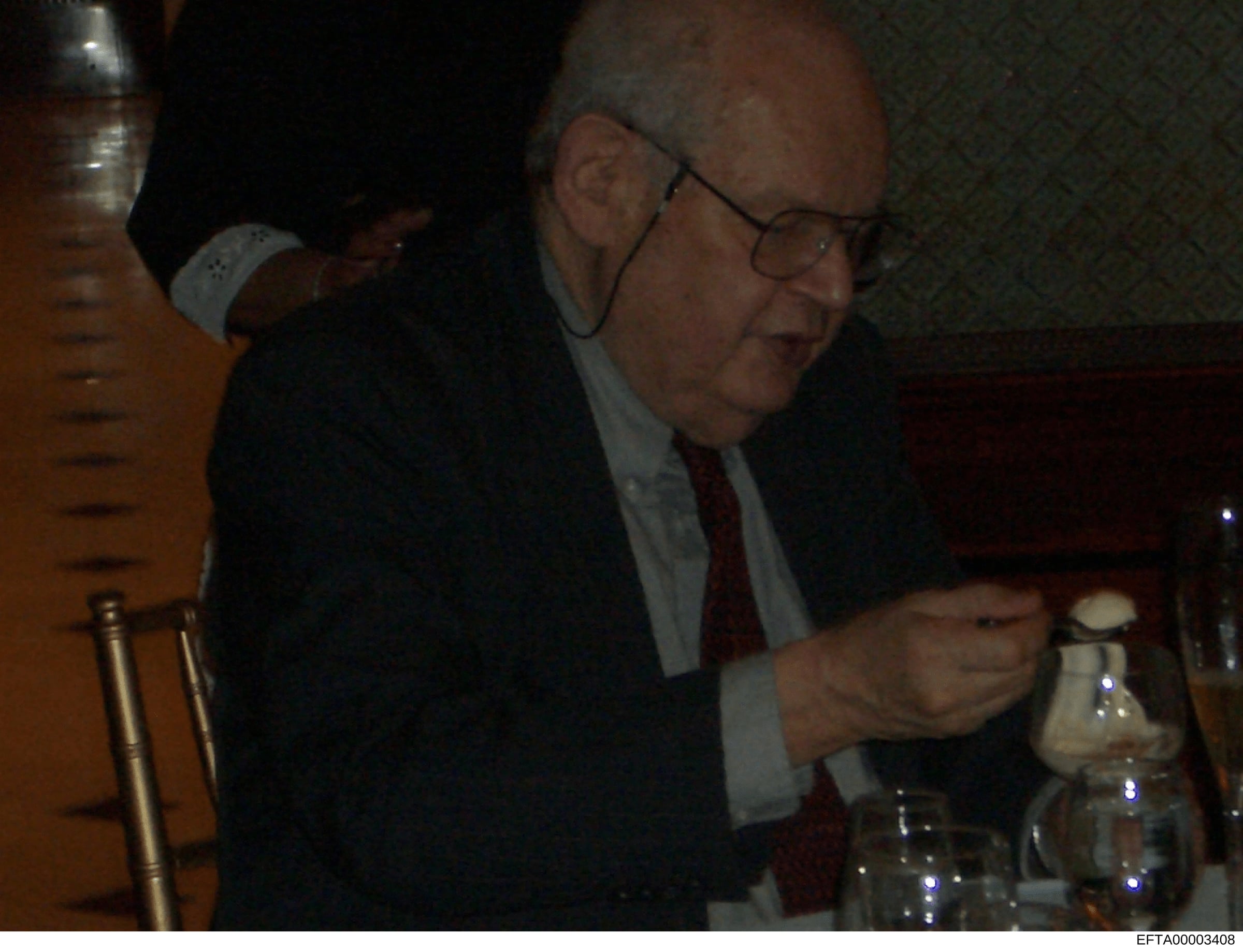 This is an evidence photograph from the Epstein investigation files showing an unidentified man wearing glasses and formal business attire (dark suit, light blue shirt, dark tie) at what appears to be an indoor social or professional gathering. The p