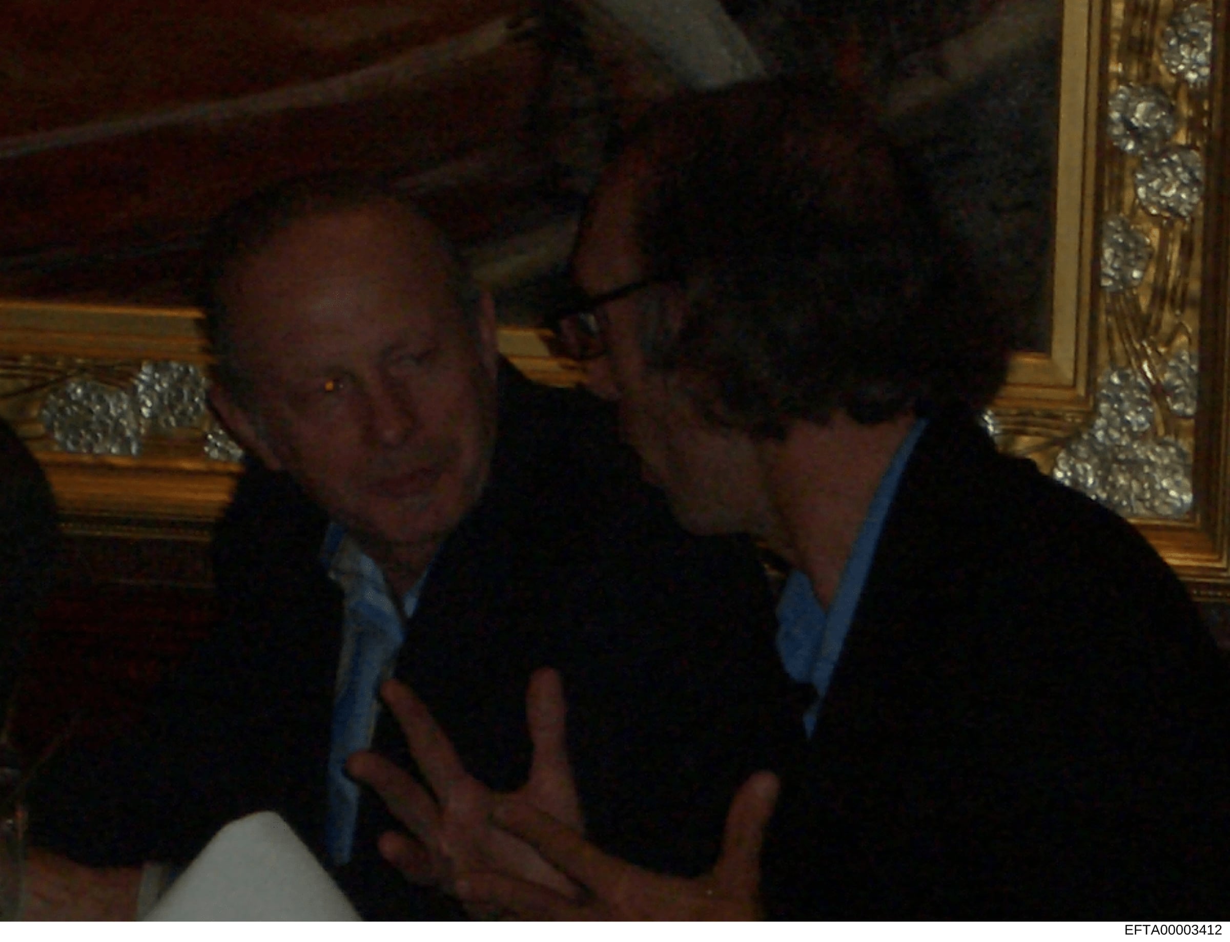 This photograph shows two men engaged in conversation in what appears to be an ornately decorated interior space. The setting features gold-trimmed frames and formal furnishings, suggesting an upscale residence. The photo quality and color characteri