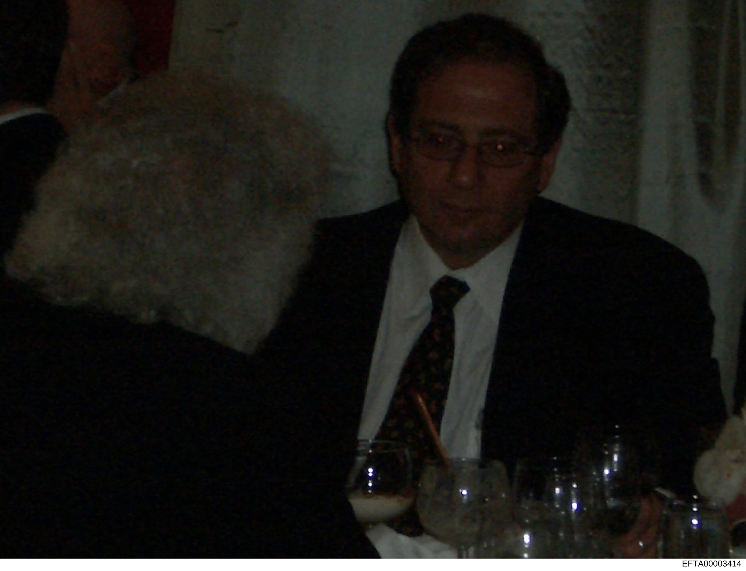 A photograph of a middle-aged man wearing glasses, a dark suit jacket, white dress shirt, and patterned tie, seated at what appears to be a formal indoor event or gathering. Water glasses are visible on the table in front of him. The image quality is