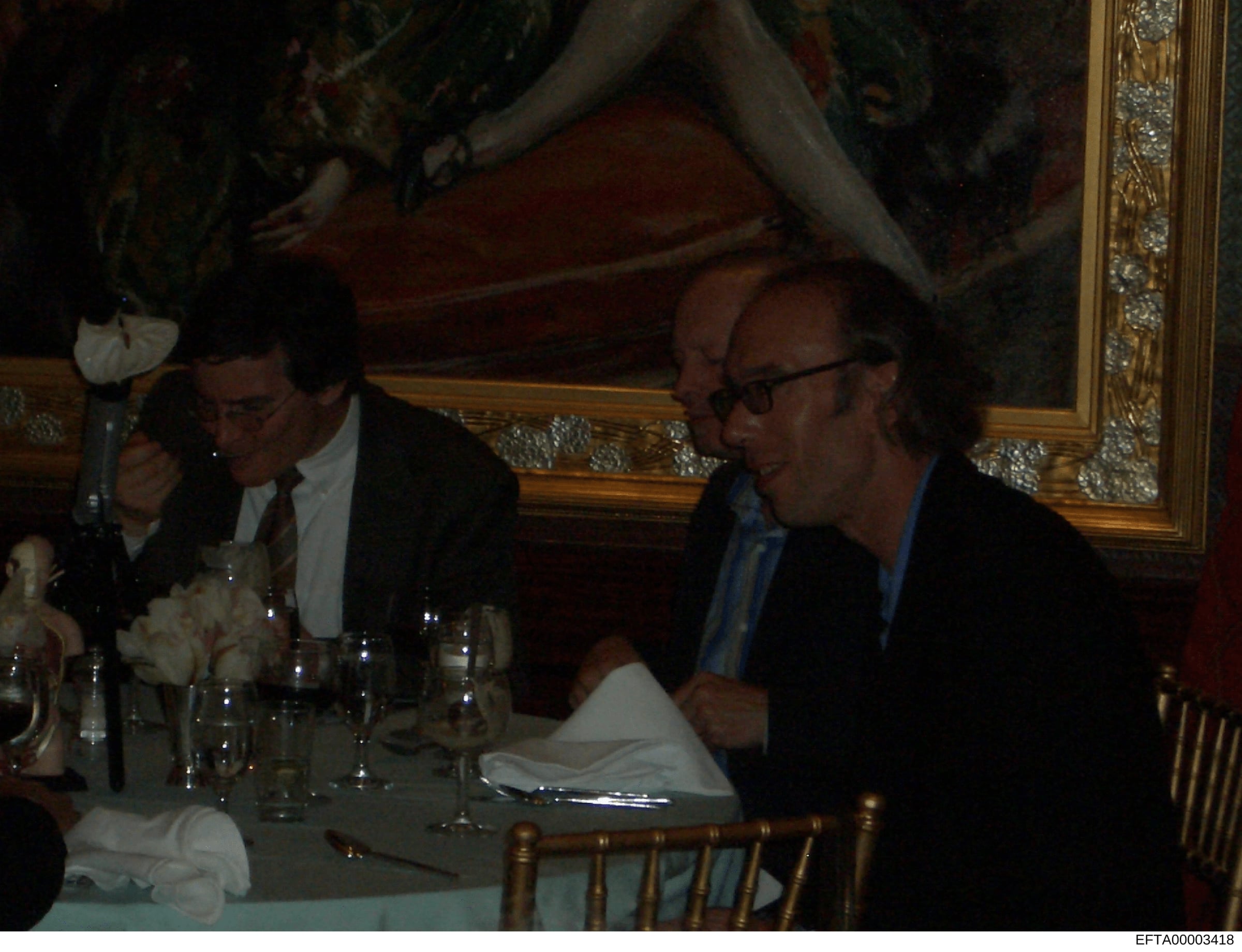 This is a photograph of what appears to be a formal dinner event in an ornately decorated room. Multiple people are seated at a dining table with place settings, glasses, and napkins. The setting suggests an upscale venue with decorative molding and